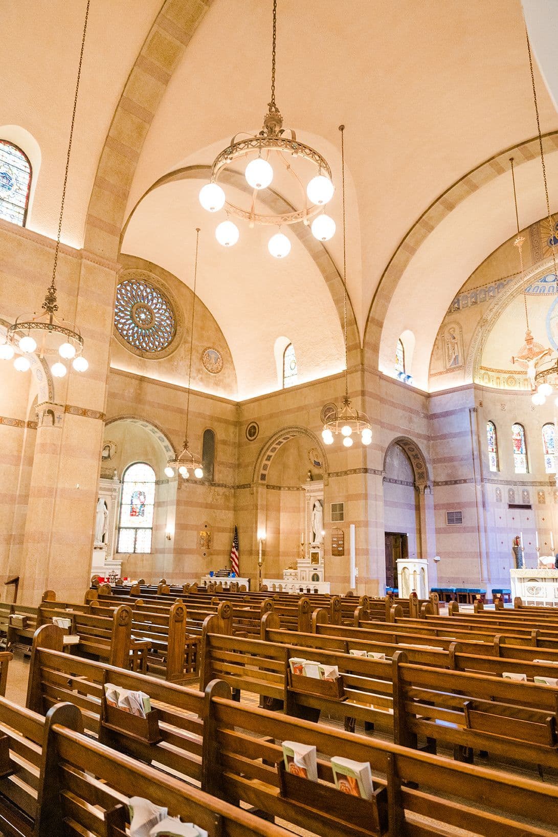 The interior of St. Mary's Catholic Church looking toward the sanctuary — stone arches, a rose window, hanging globe chandeliers, and rows of wooden pews.